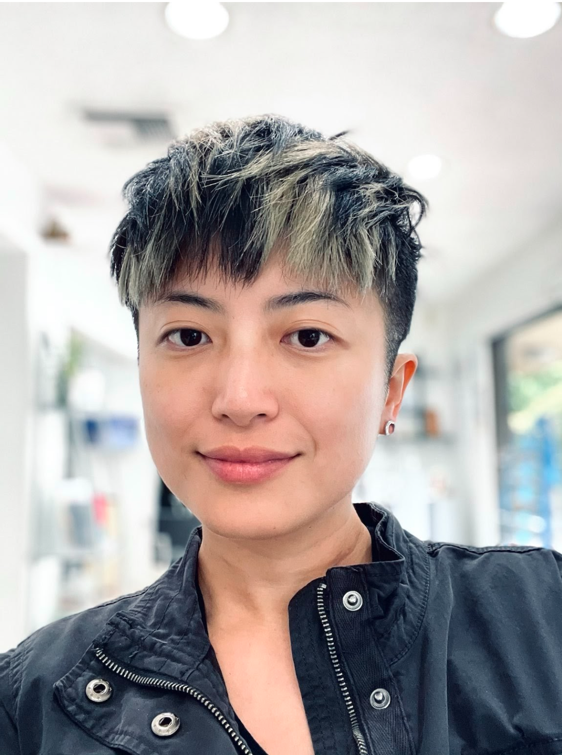  a Han-Taiwanese person with light yellow colored skin and very short hair with blond highlights stands in a sun-filled room in front of multiple shelves