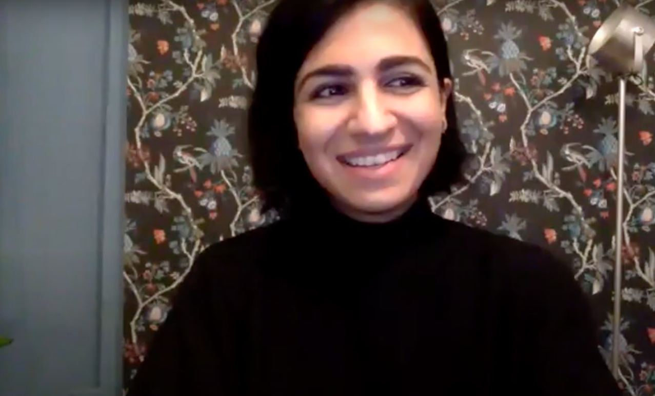 A woman with medium length dark brown hair smiles in front of a dark floral wallpaper background during a Zoom webinar.
